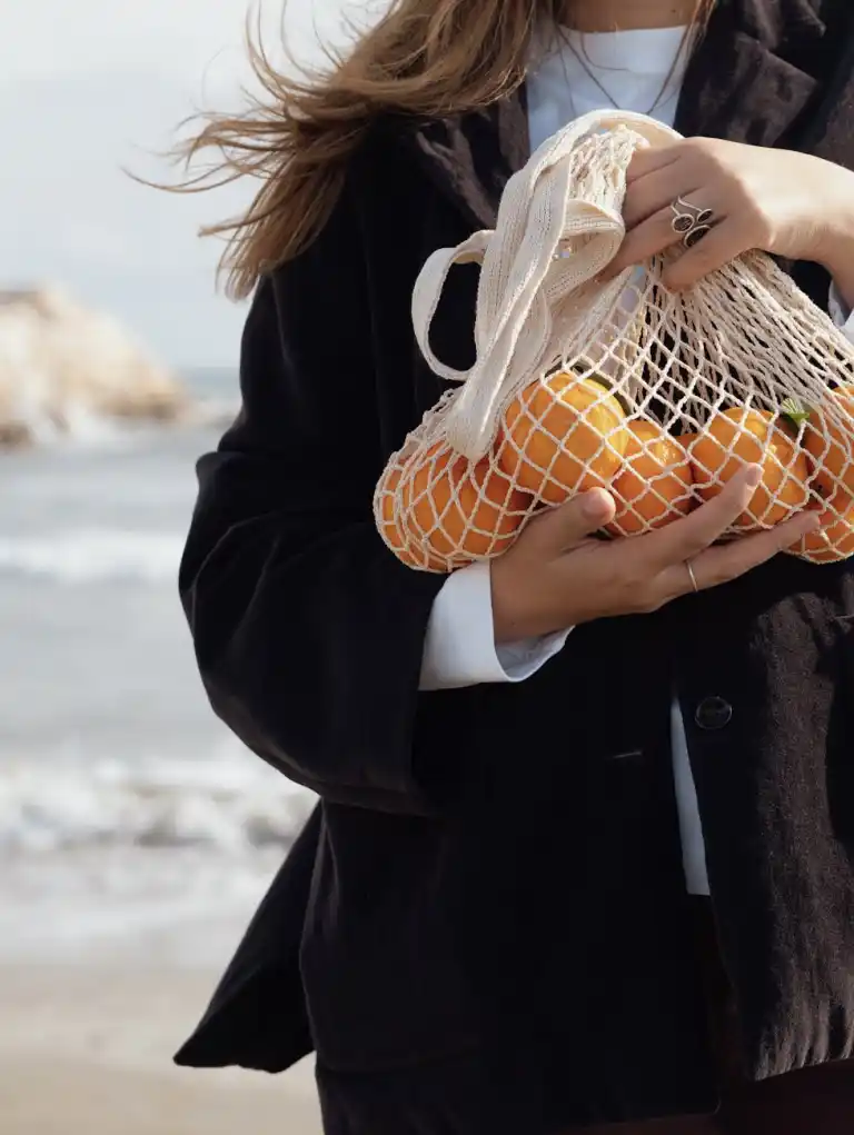 woman holding oranges in mesh bag by beach